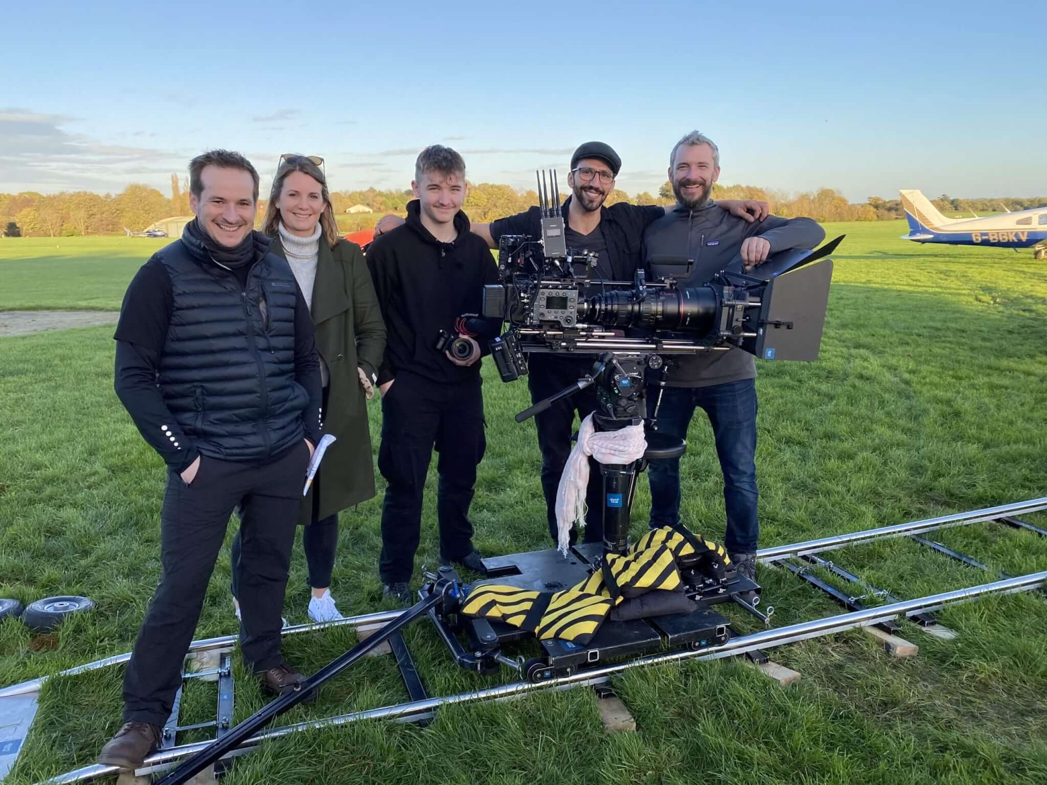 A group of five people stands together on a grassy field next to camera equipment on tracks, embodying the vibrant spirit of video production Surrey. They smile at the camera with a small airplane and trees visible in the background under a clear sky.