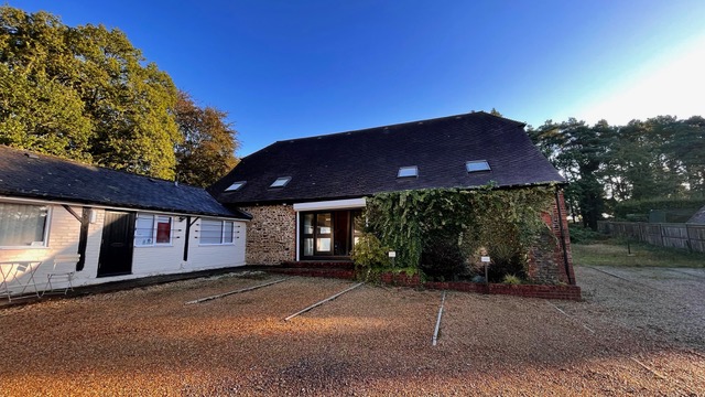 A quaint house with a steep roof and ivy-covered entrance, set against a clear blue sky. The house is adjacent to a white building with black shutters. A gravel driveway with marked parking spots is in the foreground. Trees frame the setting.
