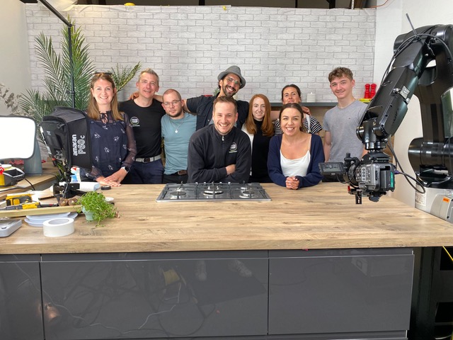 A group of ten people smiling and posing behind a kitchen counter with a gas stovetop. There are filming equipment and plants around. The background features a light gray brick wall.