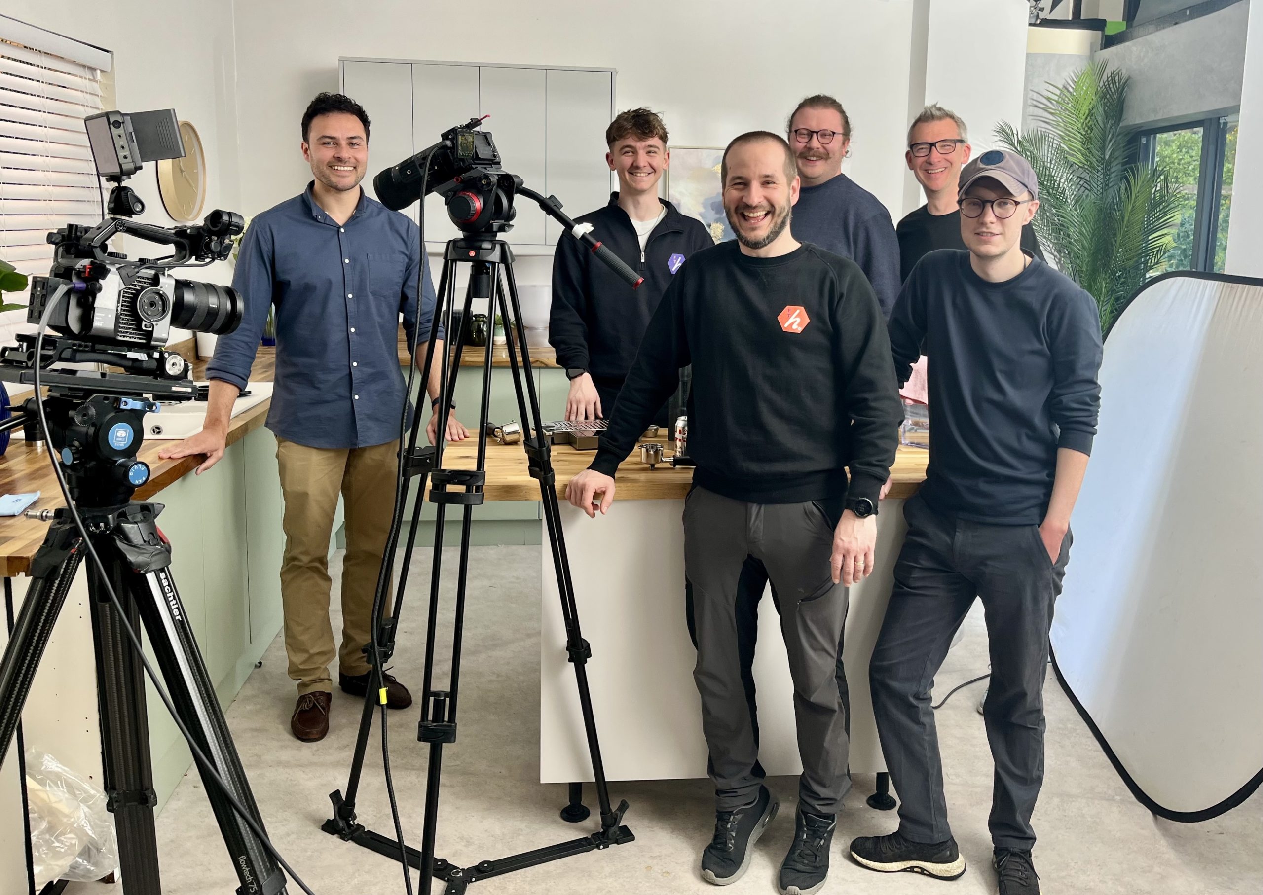 Group of six men smiling in a studio, with camera equipment set up around them. They stand near a table with various items on it. The setting is bright, with plants and lighting equipment in the background.