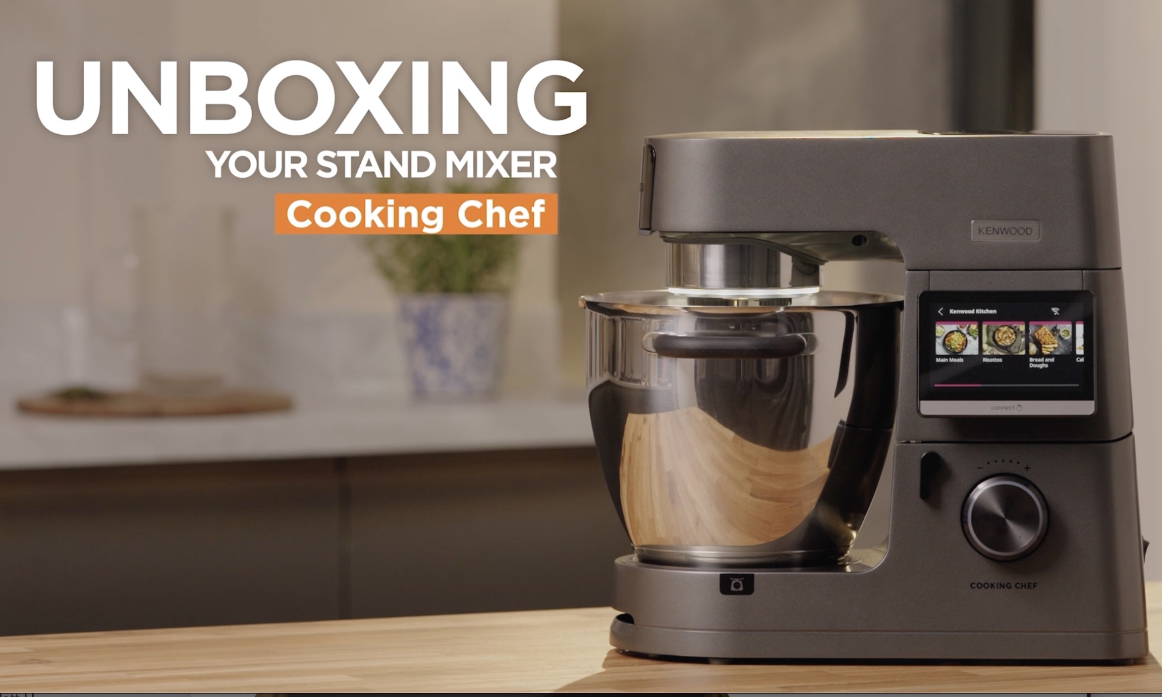 A sleek, metallic stand mixer is displayed on a kitchen counter. The screen shows digital features, and text on the image reads Unboxing Your Stand Mixer Cooking Chef. A vase with a plant is in the blurred background.