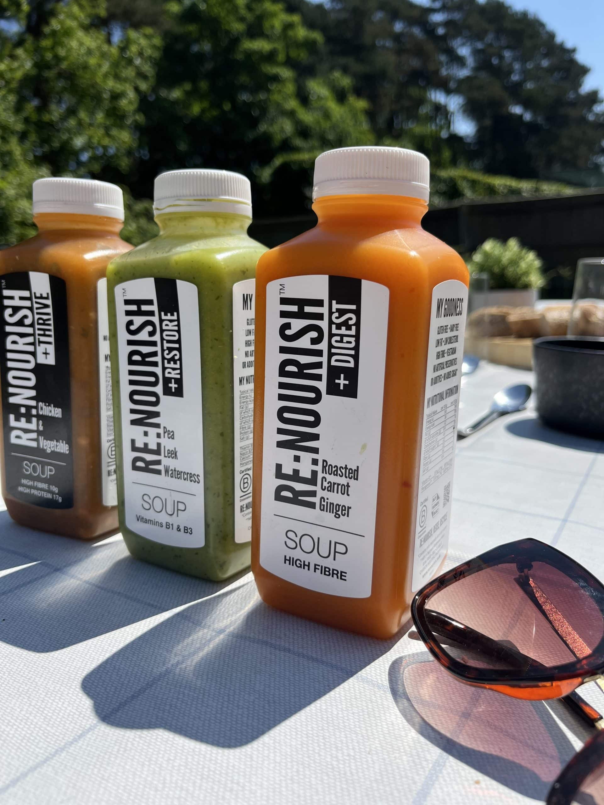 Three bottles of RE:NOURISH soup sit on an outdoor table. Flavors include Chicken & Veg, Pea Leek Watercress, and Roasted Carrot Ginger. Sunglasses and sunlight add to the summer picnic atmosphere.