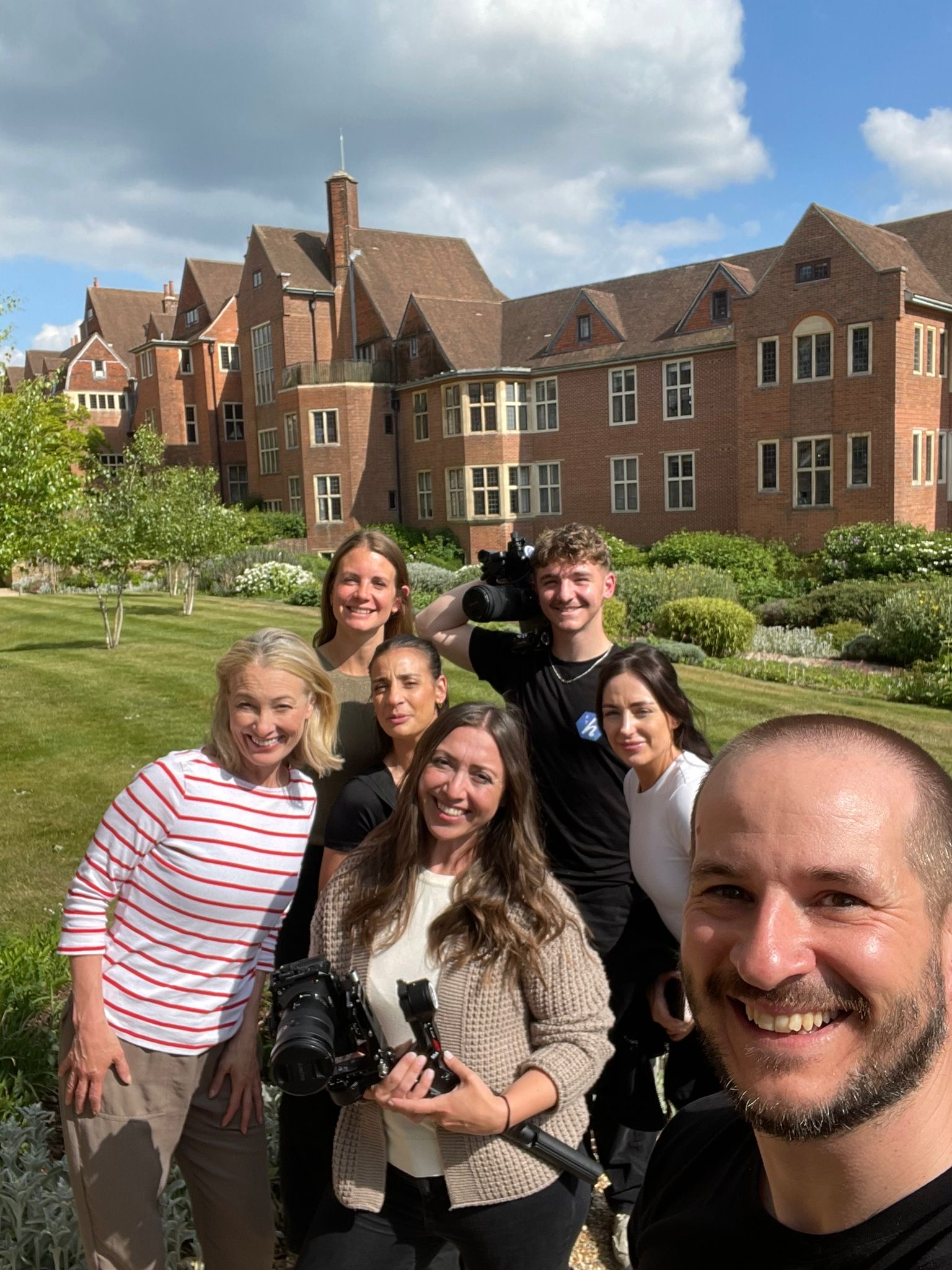 A group of seven people, some holding camera equipment, smile for a selfie outside a large brick building with gabled roofs, blending city and country vibes amid a lush garden under a partly cloudy sky.