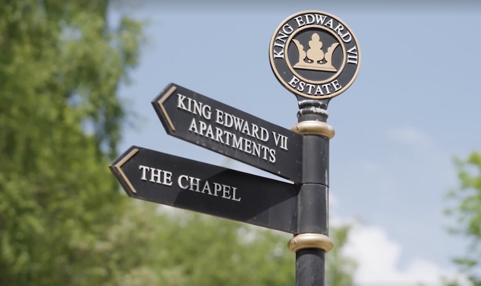 A black metal signpost with gold accents points to King Edward VII Apartments and The Chapel, beneath a round King Edward VII Estate sign. Green trees and a blue sky highlight the scene, perfect for captivating drone filming.