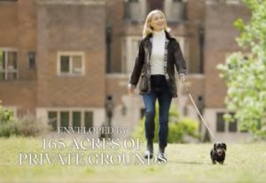 A woman in casual clothes walks a small dog on a leash across a grassy area, with a large brick building in the background. Text reads, Enveloped by 165 acres of private grounds—a perfect blend of city and country living.