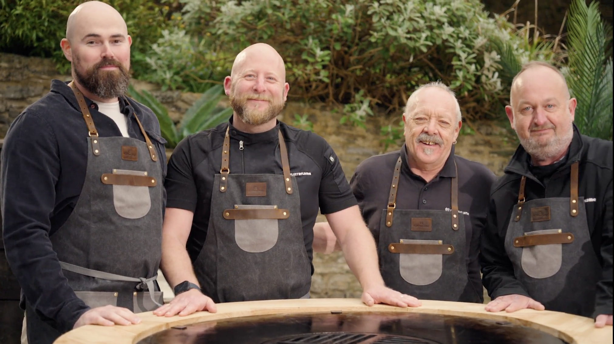 Four men wearing matching aprons stand outdoors by a circular wooden grill, smiling at the camera, with greenery and a stone wall in the background.