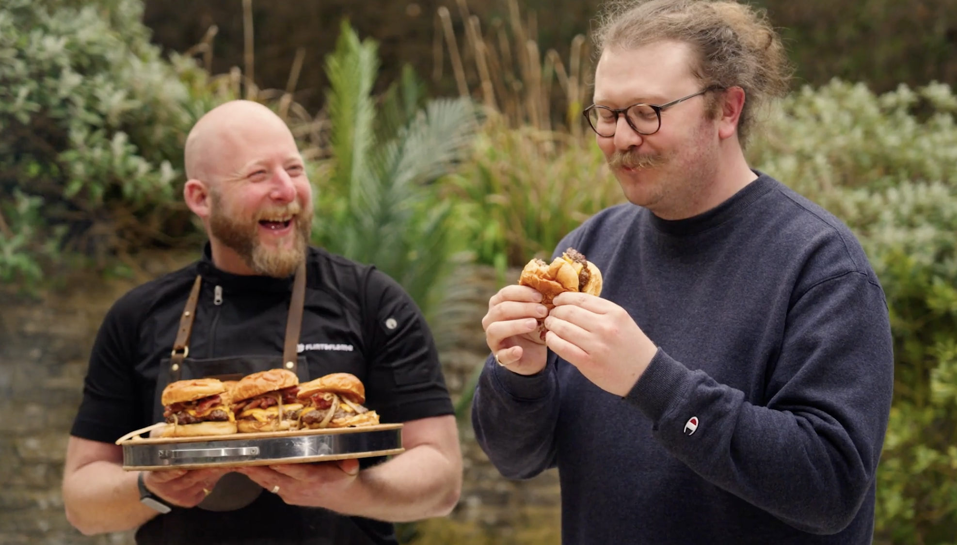 Two men stand outdoors; one is holding a tray of burgers and laughing, while the other smiles and eats a burger. There is greenery visible in the background.