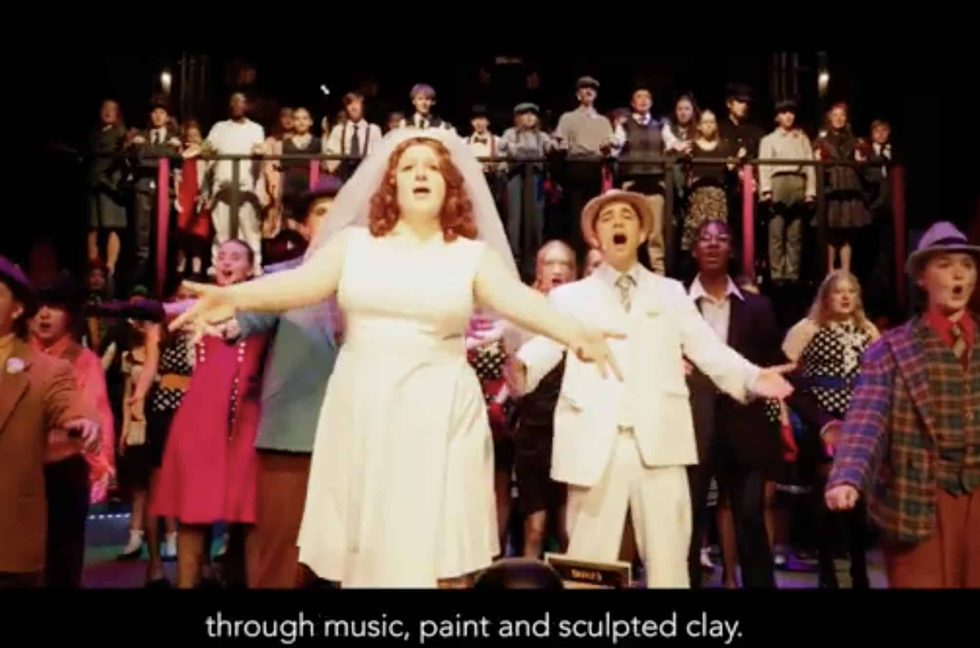 A group of performers on stage sing passionately, with a woman in a white dress and veil and a man in a white suit at the front, as a large chorus stands behind them—an inspiring scene from King's InterHigh Online School.