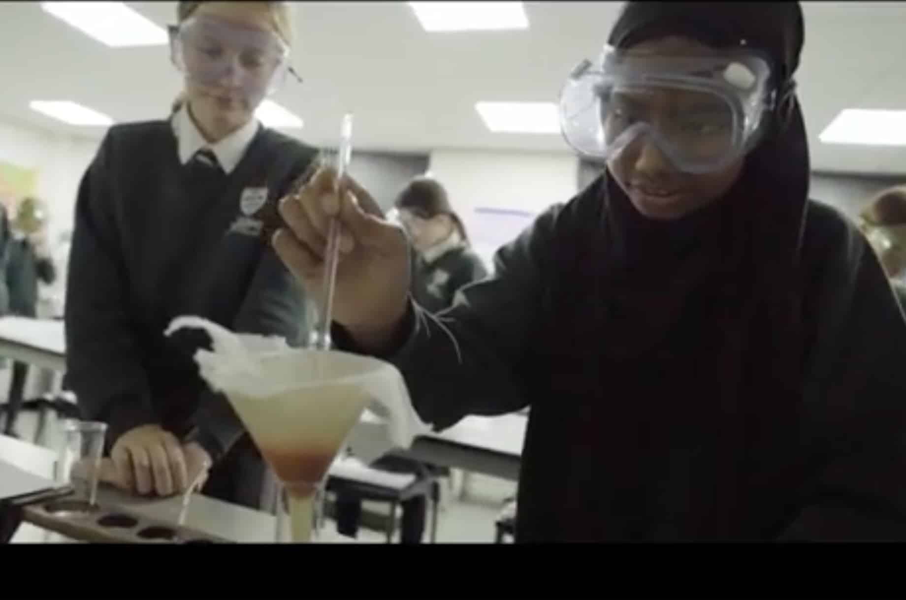 Two students in King's InterHigh Online School uniforms and safety goggles conduct a science experiment in a classroom. One pours liquid with a pipette into a funnel lined with filter paper as the other observes closely.