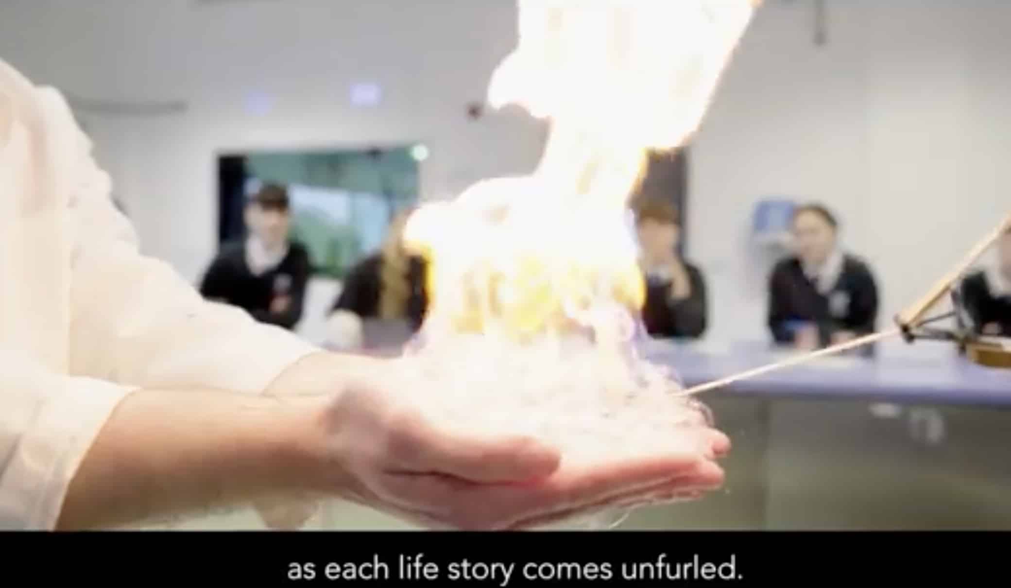A person holds a controlled flame in their hand during a science demonstration at King’s InterHigh Online School, with several people watching in the background. Text at the bottom reads, as each life story comes unfurled.