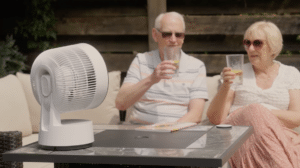 An elderly couple sits outdoors on a patio sofa, wearing sunglasses and holding drinks with lemon slices, as a white electric fan cools them nearby. They enjoy relaxing after watching Outdoor Cooking Videos together.