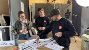 Three people stand around a table covered with papers, discussing documents. One person points at a sheet while the others watch, all smiling. Studio equipment and a large light suggest they might be planning their next Outdoor Cooking Videos.
