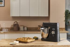 A black stand mixer sits on a wooden kitchen worktop next to a freshly baked rectangular cake topped with seeds. Small bowls with ingredients are nearby, creating an inviting scene perfect for fans of Outdoor Cooking Videos.
