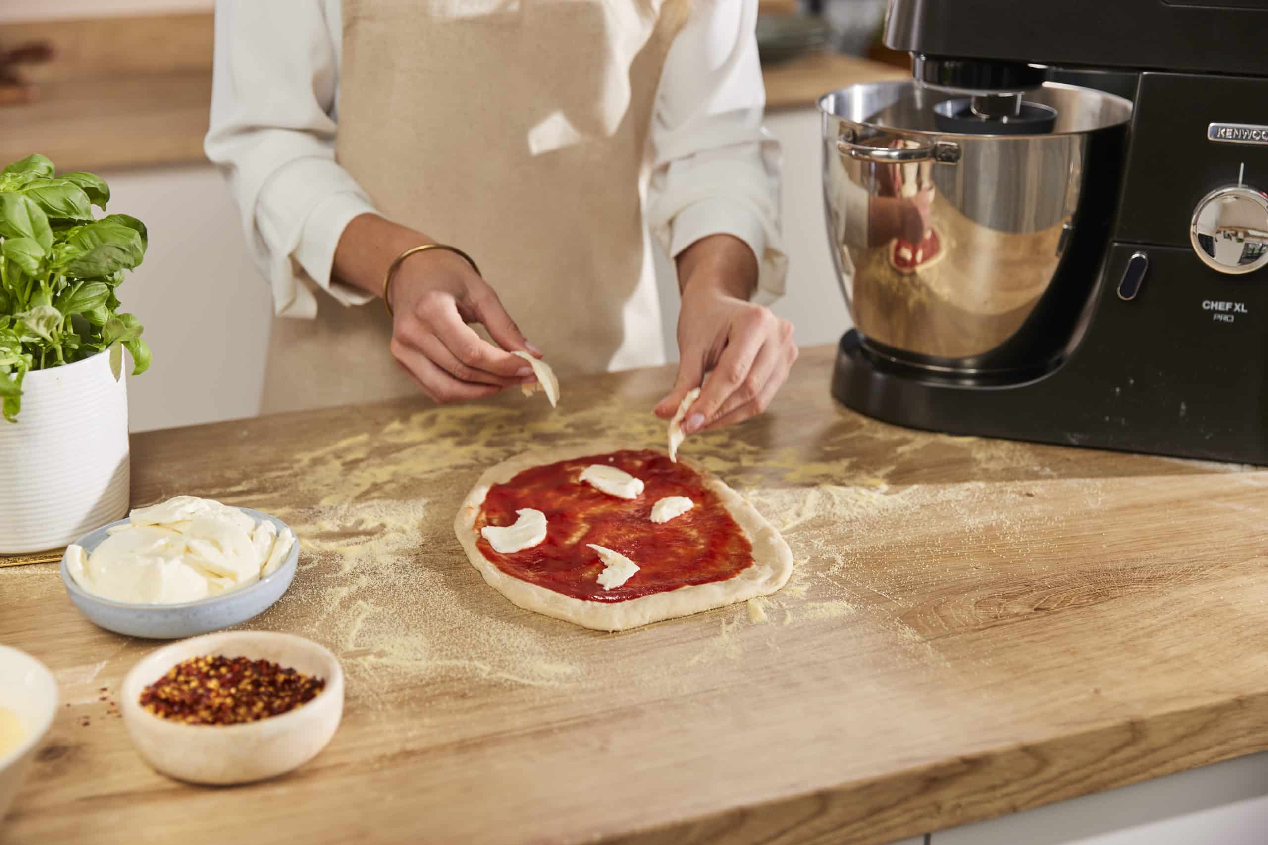 A person in an apron adds pieces of cheese to a pizza with tomato sauce on a floured wooden counter, surrounded by basil, mozzarella, and chilli flakes, next to a stand mixer—perfect for fans of Outdoor Cooking Videos.