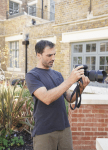 A man in a black T-shirt examines a camera outdoors in front of a brick building with large windows and plants, preparing to film outdoor cooking videos.