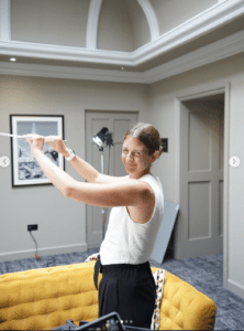 A woman smiling and stretching her arms whilst holding a white rod, standing beside a yellow sofa in a modern, well-lit room with grey walls and framed artwork, perhaps inspired after watching some Outdoor Cooking Videos.