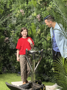 A young girl in a red shirt and leopard print trousers smiles whilst standing next to a man in a blue shirt in front of a lush, green, plant-filled wall. A shiny, metallic object for outdoor cooking videos sits in front of them.