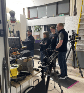 Four people stand in a studio, focused on a monitor with camera equipment and cables around them, reviewing their latest Outdoor Cooking Videos as one person gestures towards the screen while others watch attentively.