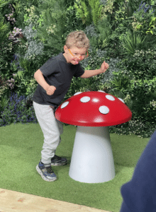 A young boy wearing a black shirt, grey tracksuit bottoms, and orange sunglasses playfully poses next to a large, red-and-white spotted toadstool sculpture, with lush green foliage in the background—perfect inspiration for creative outdoor cookery videos.