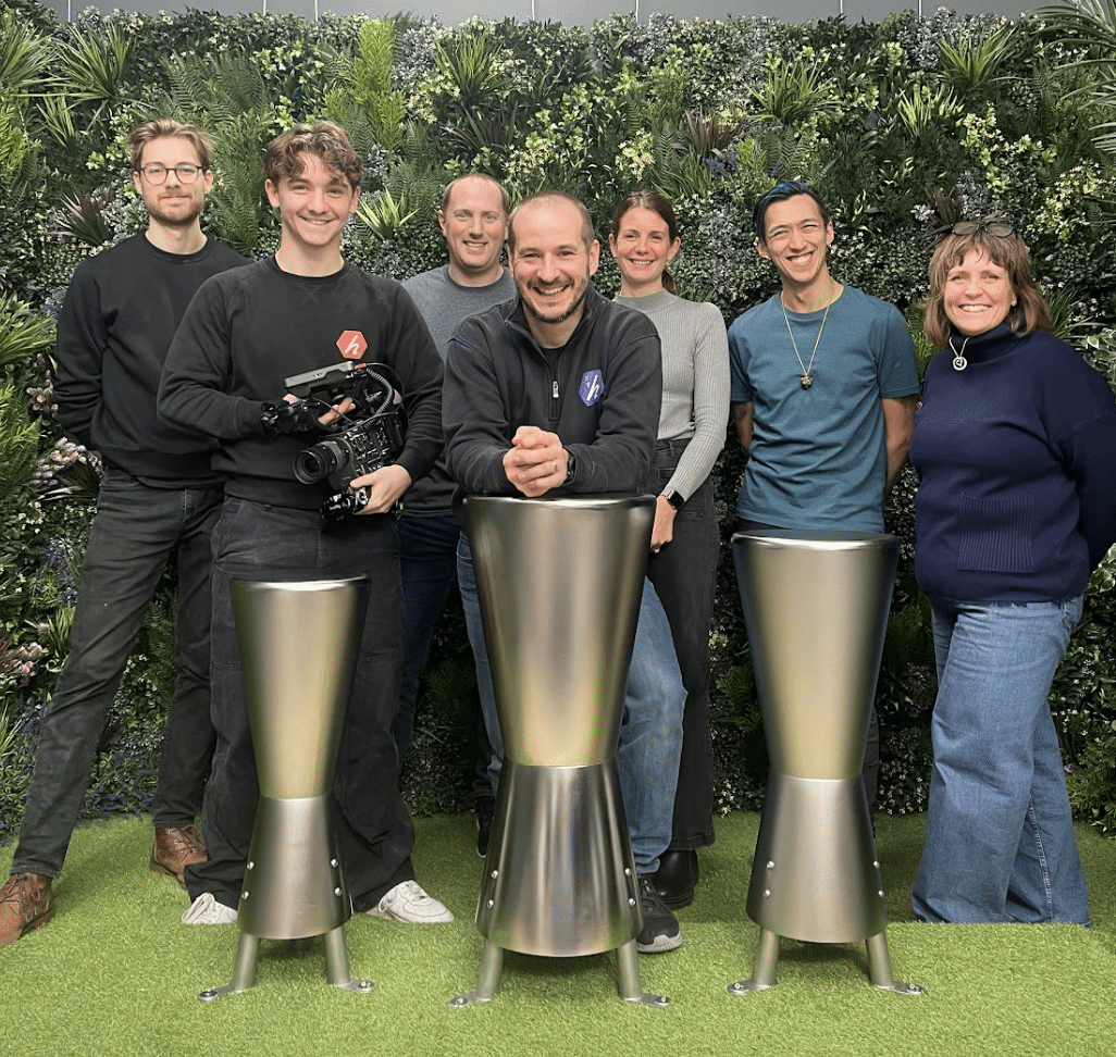 Seven people stand and smile in front of a green leafy backdrop, three metallic rocket-like objects on artificial grass hinting at Outdoor Cooking Videos. One person holds a camera, while the cheerful group appears casual and relaxed.