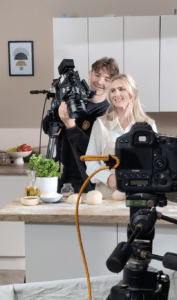 A smiling woman stands in a kitchen next to a cameraman holding a large camera, ready to film. Another camera sits in the foreground, and ingredients like dough, basil, and olive oil hint at delicious recipes for outdoor cooking videos.