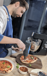 A man in an apron uses a heat gun to finish a homemade pizza on a wooden board in a modern kitchen, with another pizza, a bowl of chilli flakes, and a stand mixer visible—perfect inspiration for Outdoor Cooking Videos.