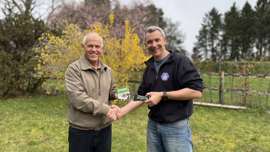 Two men stand outdoors shaking hands and smiling, each holding a pack labelled “FOOD.” In a grassy garden with blooming trees behind them, they celebrate together—captured perfectly by video production Surrey.